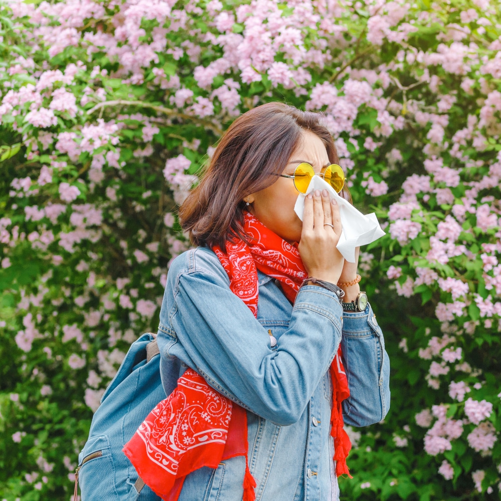 a woman standing alongside shrubbery blows her nose